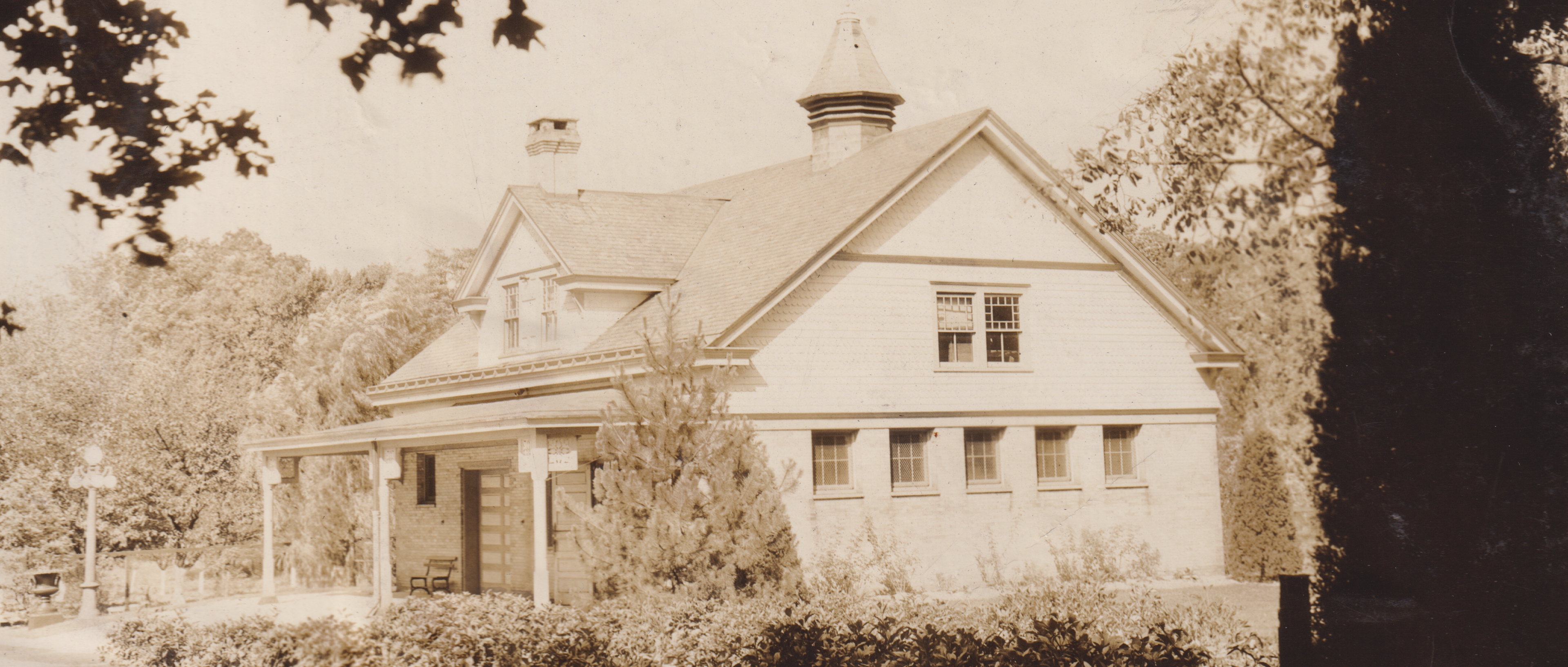 A black-and-white photograph of one of the buildings on the Haddonfield campus of the Bancroft School. The building is a small light-colored carriage house with a pointed tower on the roof, surrounded by trees and bushes.