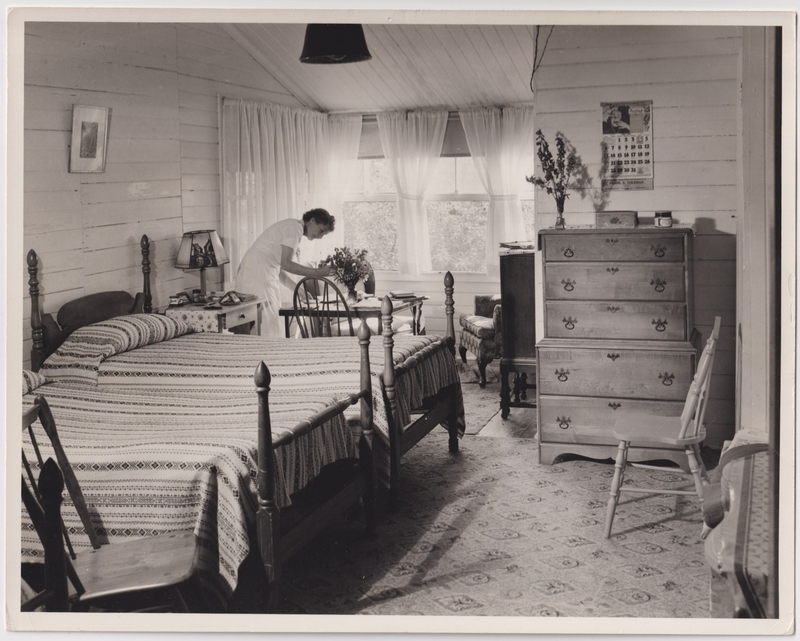 A black-and-white photograph of a dormitory with two beds and a table near a window, with a staff member seated at the table.